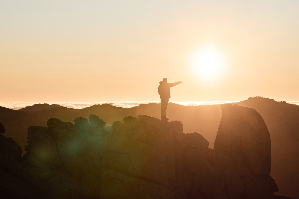 Person Standing On Top Of Rocks Pointing To The Sun at Sunrise