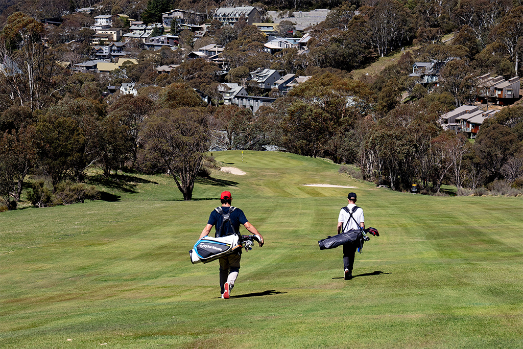 two golfers walk down the green way holding golf bags