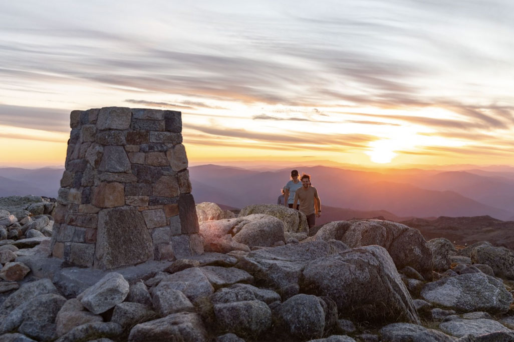 Hikers walk towards Mount Kosciuszko Summit marker with sunsetting in back ground