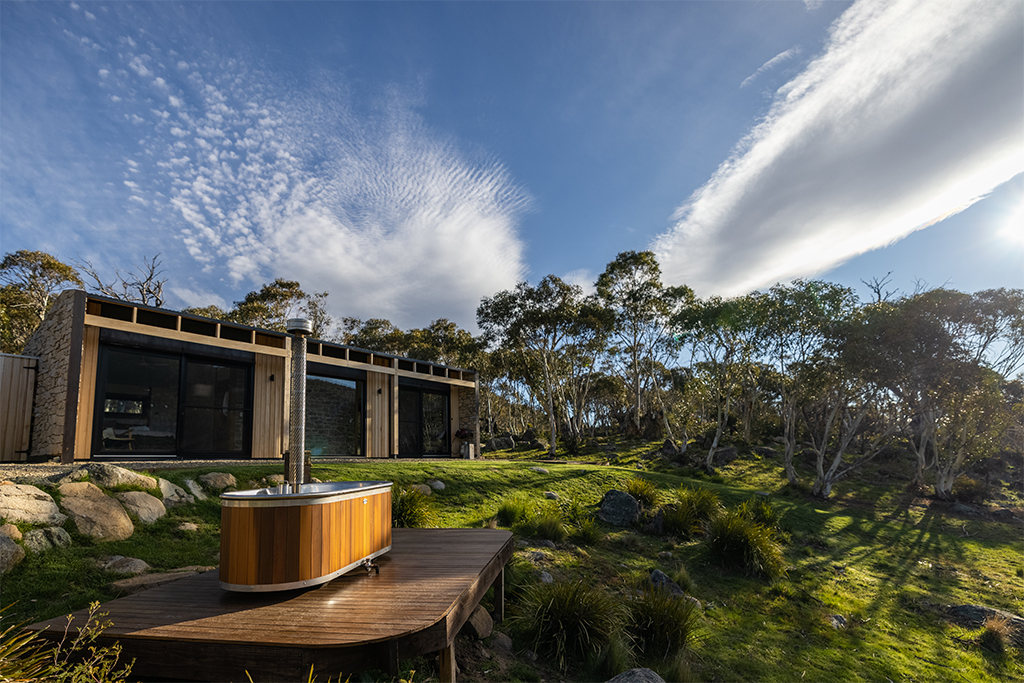 Timber hot tub sits on deck outside timber and stone cabin